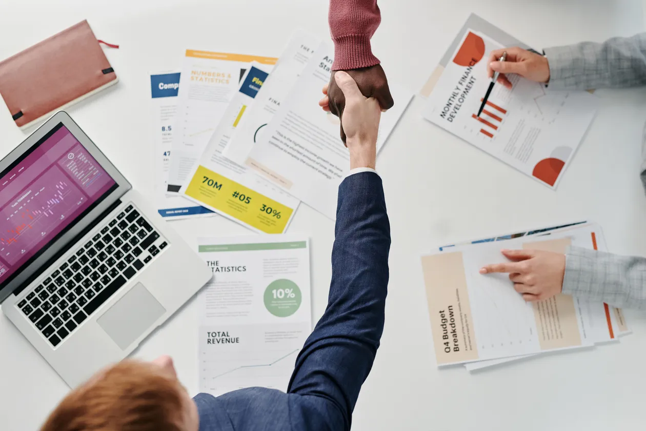 Overhead view of a handshake across a desk covered with a laptop, statistics documents, monthly finance charts, and budget breakdown infographics