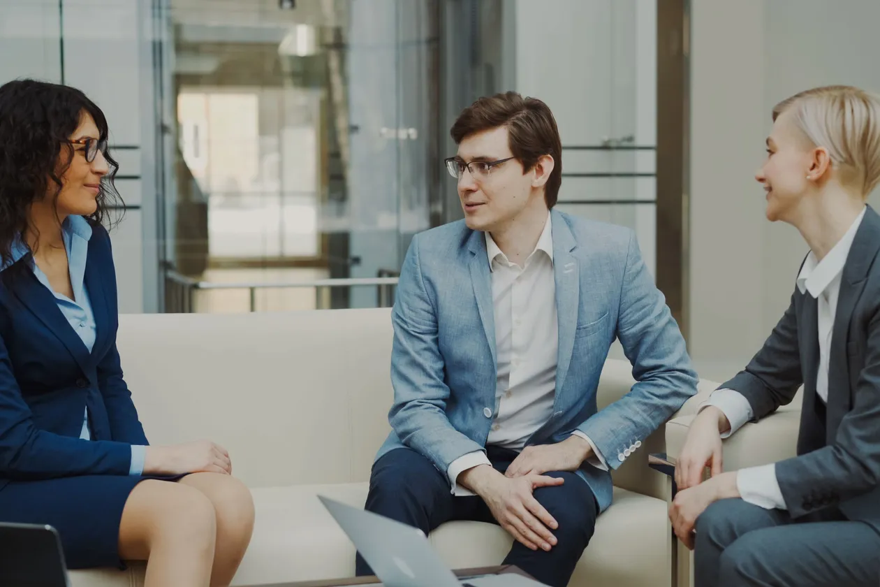Three professionals in business attire seated in a modern office having a discussion, with a laptop on the low table beside them