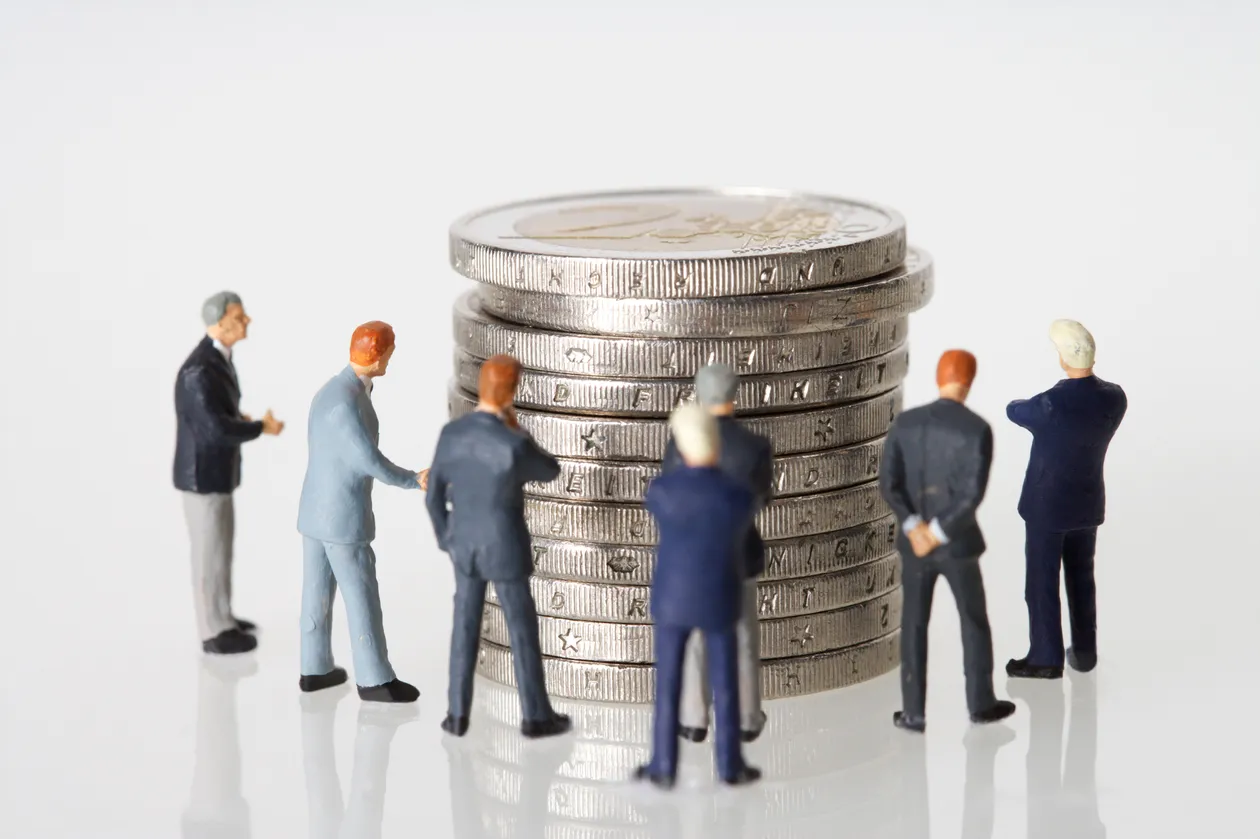 Miniature figurines of business people gathered around a tall stack of silver coins, symbolizing negotiation