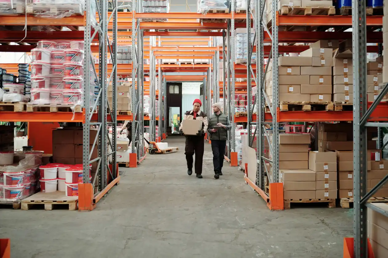 Two warehouse workers walking down an aisle between tall metal shelves stacked with cardboard boxes and pallets