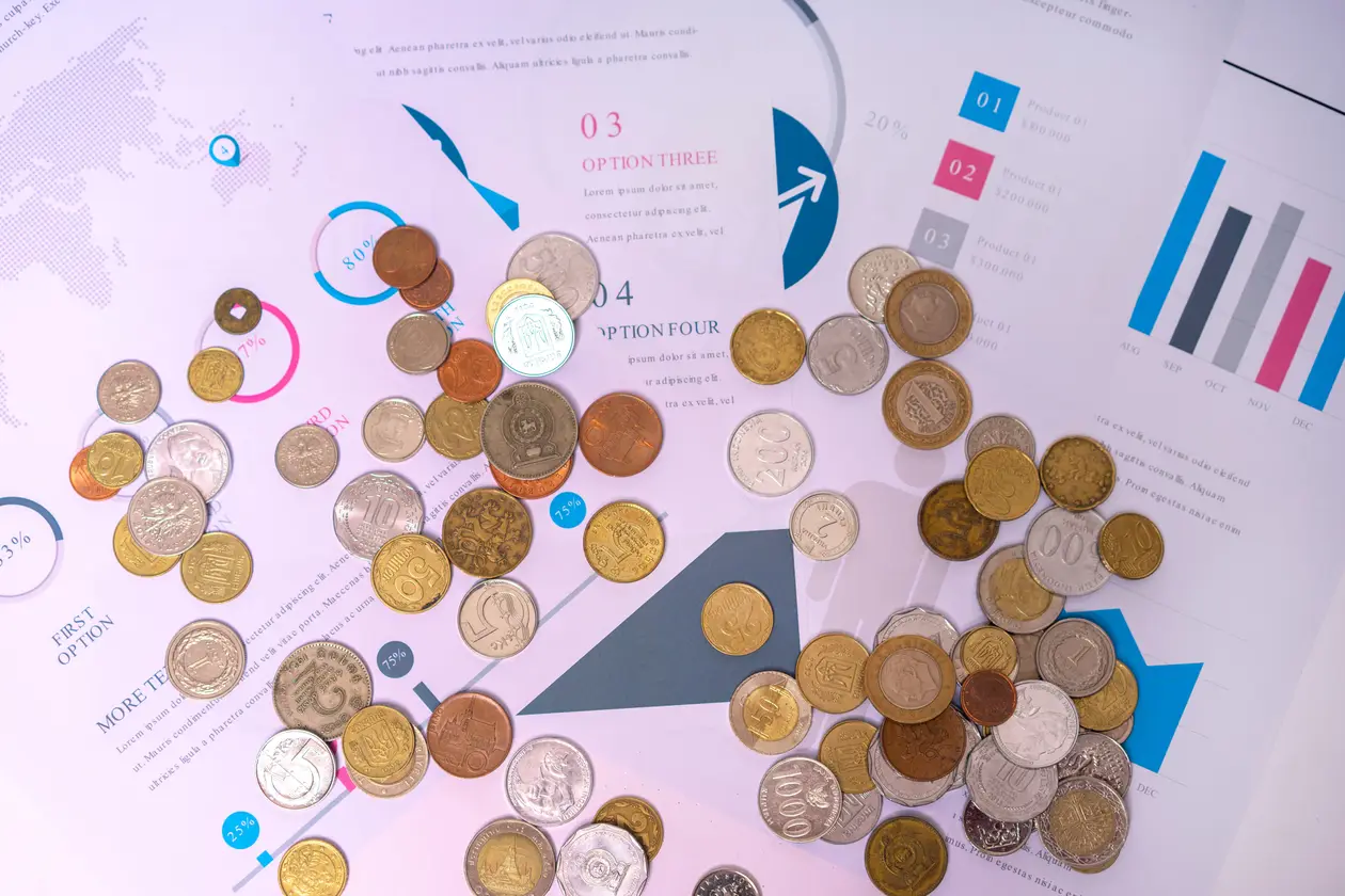 Assorted coins scattered across printed business reports and infographic pages showing charts and options