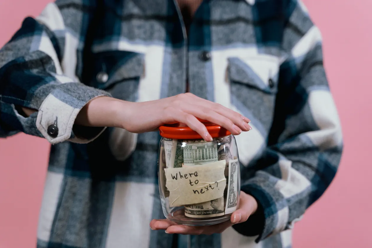 Person in a blue plaid flannel shirt holding a glass jar of cash labeled 'where to next?'