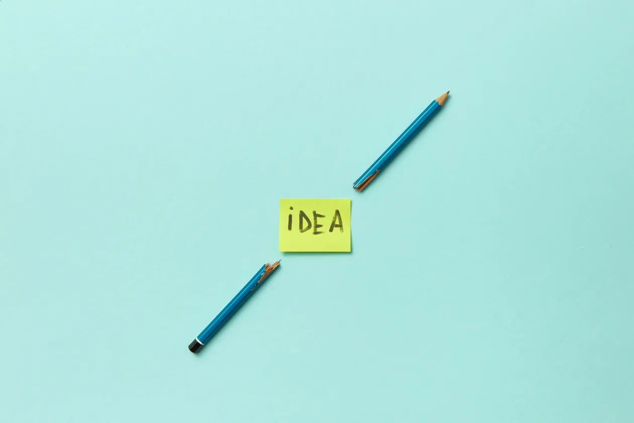 Broken blue pencils flanking a yellow sticky note that says 'iDEA'