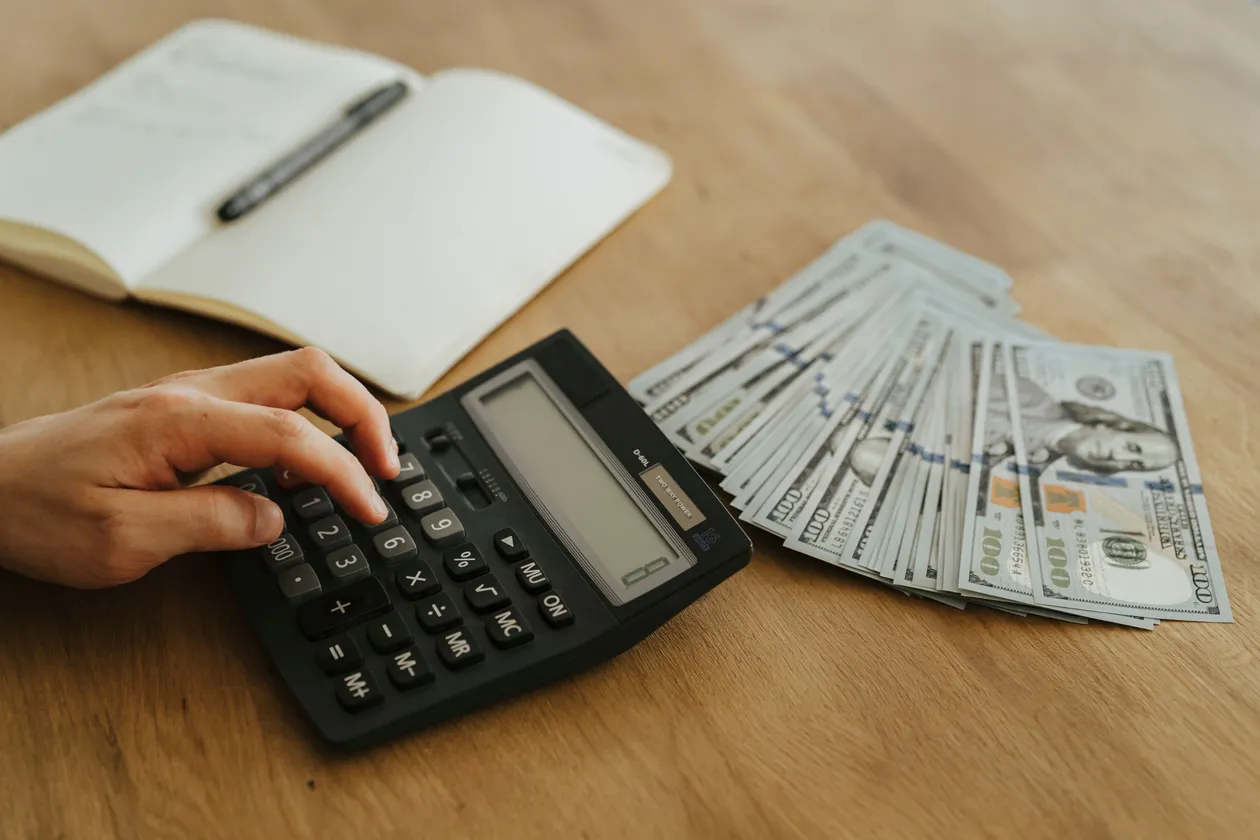 Person's hand pressing keys on a black desk calculator beside a fan of hundred-dollar bills and an open notebook