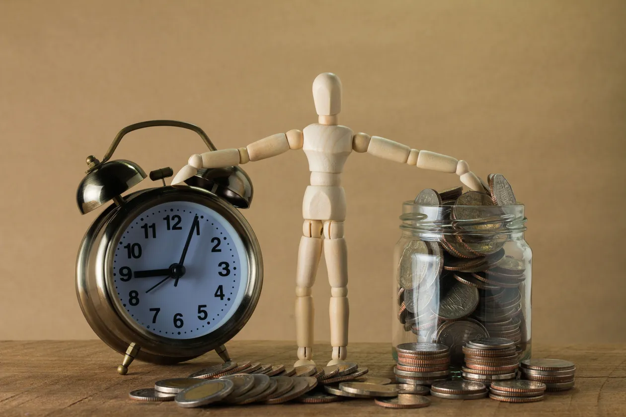Wooden doll figurine standing between a vintage alarm clock and a jar overflowing with silver coins on a rustic tabletop
