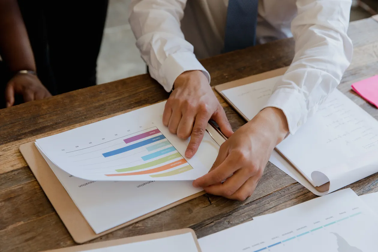 Person in a white shirt pointing at a colorful bar chart on a document at a wooden desk