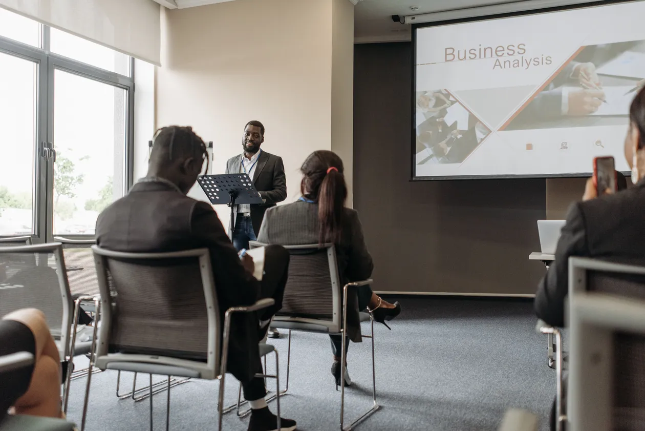 Speaker at a lectern presenting business analysis slides to a small audience seated in a conference room
