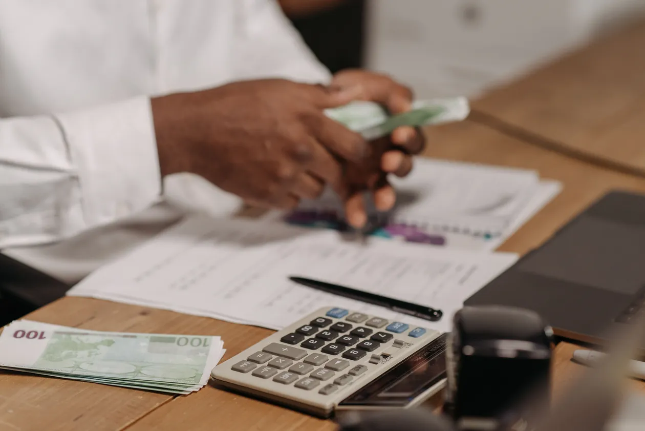 Person in a white shirt counting banknotes at a wooden desk cluttered with financial documents and a calculator