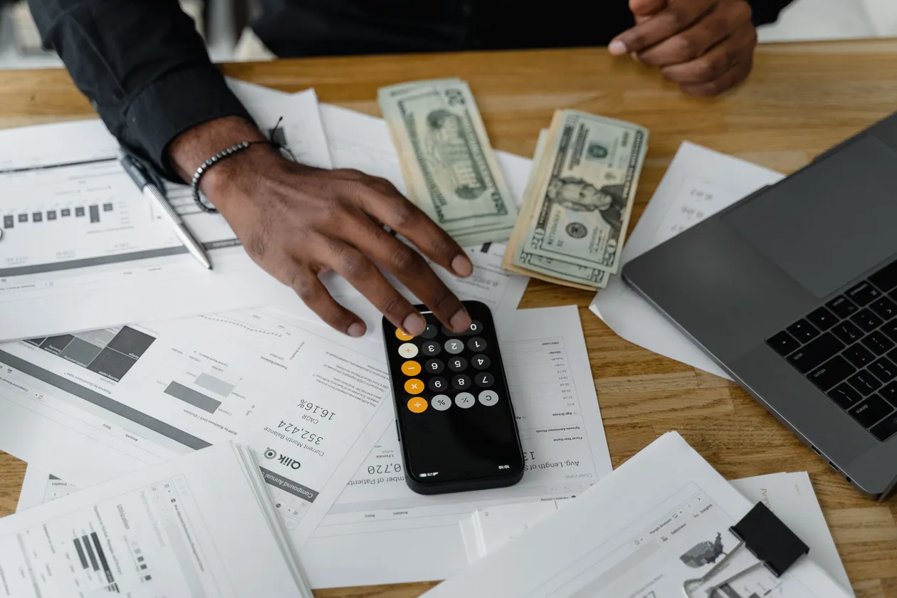 Business owner using a calculator surrounded by financial documents, cash, and a laptop on a wooden desk