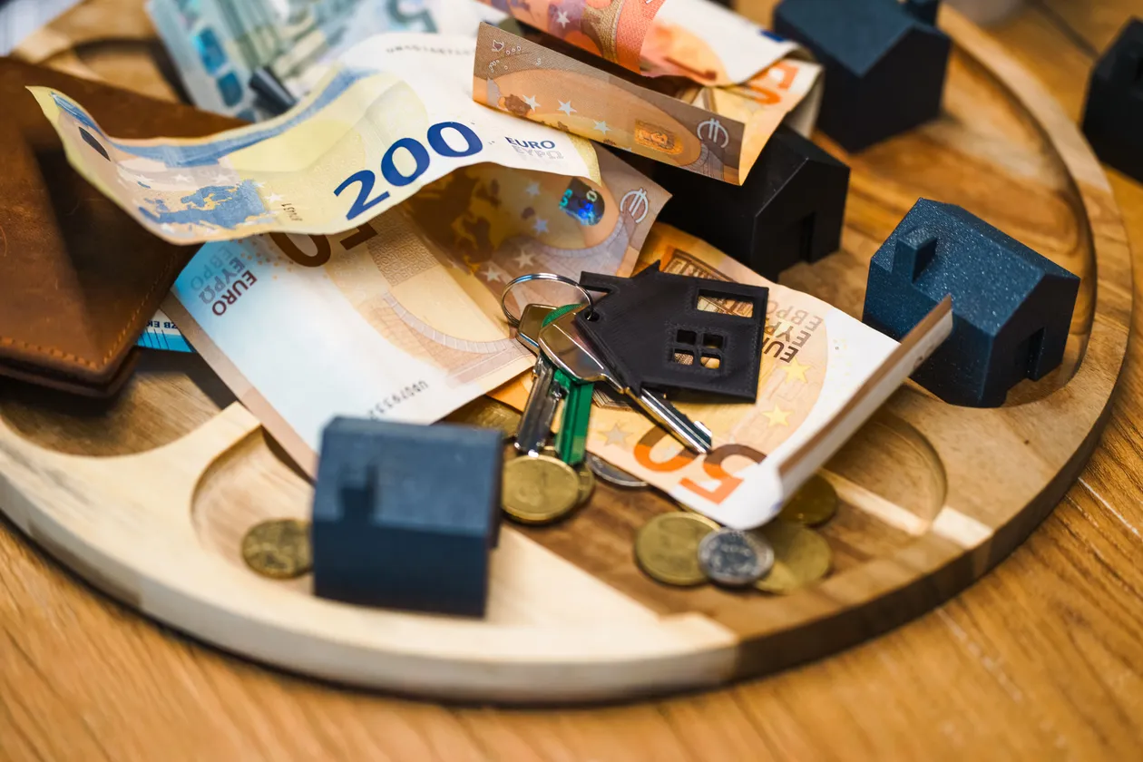 Close-up of Euro banknotes, coins, keys, and small dark house-shaped miniatures scattered on a round wooden tray