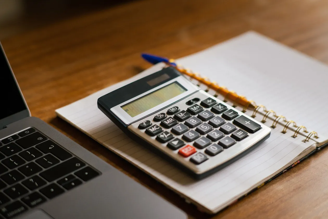 Calculator resting on a spiral notebook beside a laptop and pen on a wooden desk, representing financial oversight