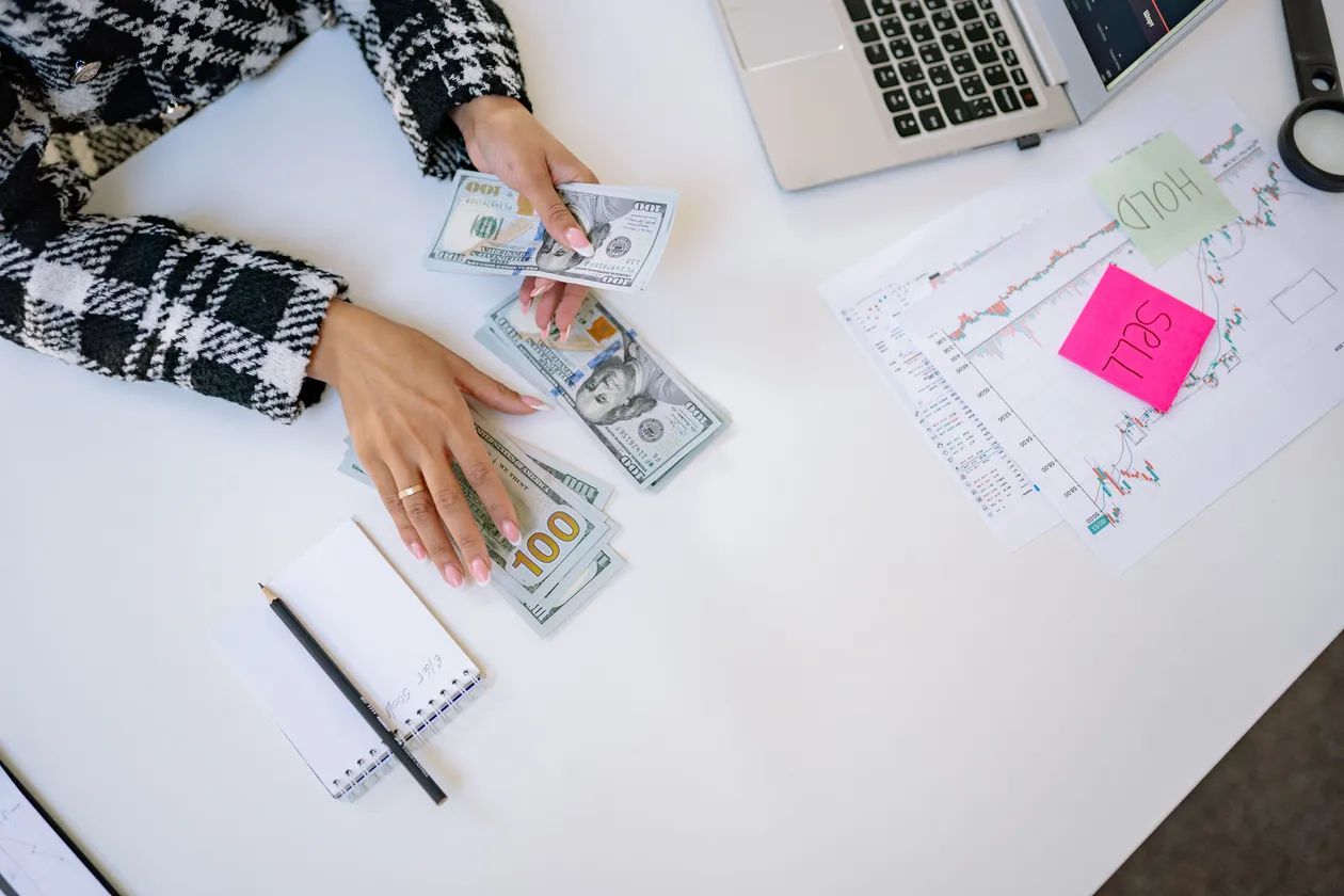 Overhead view of a person counting hundred-dollar bills at a desk with a notepad and market chart