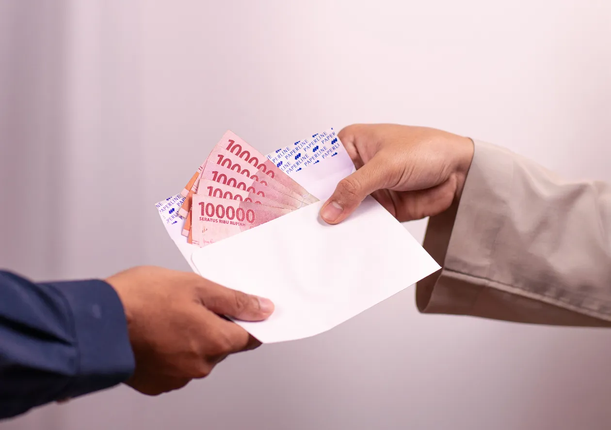 Two hands exchanging a white envelope filled with Indonesian one-hundred-thousand rupiah banknotes